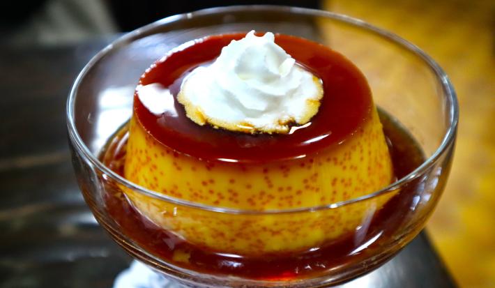 Fotografia de um mini pudim de café dentro de uma taça pequena de vidro com a calda de caramelo. Por cima do pudim tem um pouco de chantilly.