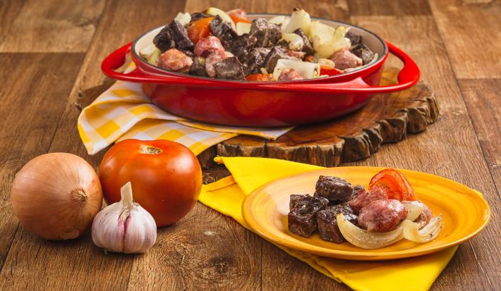 Foto de uma bancada de madeira. Nela há uma travessa vermelha com pedaços de carnes, linguiça e legumes, uma cebola, alho e um tomate na bancada e um prato amarelo com mais um pouco da receita de churrasco.