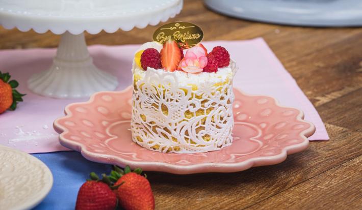 Image da receita de Mini Bolo Bem-Casado com Renda de Açúcar da Beca Milano, sobre um prato rosa, em uma bancada de madeira, decorado com frutas vermelhas e uma plaquinha com os dizeres Beca Milano em dourado