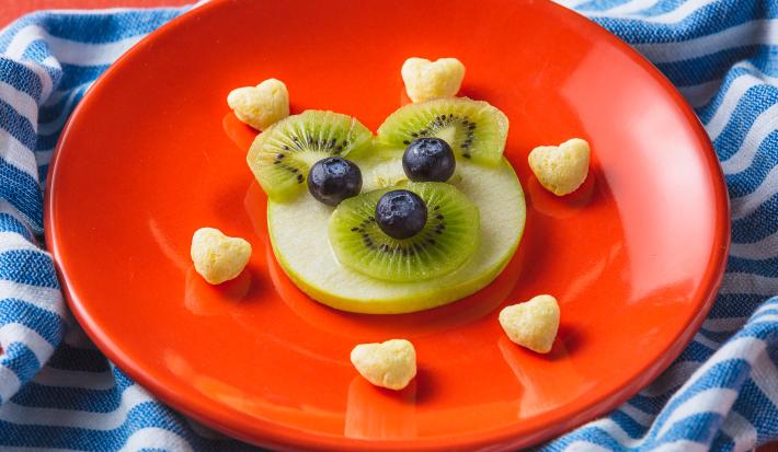 Fotografia em tons de laranja em uma bancada com paninho com listras azuis. Ao centro, um prato laranja redondo com um ursinho feito de maçã, kiwi e mirtilo e acompanhado por biscoitinhos em formato de coração. Ao lado, uma colher laranja e uma embalagem.