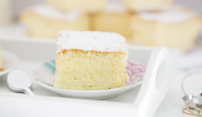Fotografia em tons claros de um bolo de massa de pão de ló com açúcar de confeiteiro em cima, que está sobre um prato pequeno branco, com desenhos em rosa e azul. Ao fundo, mais pedaços do bolo, e ao lado do prato, colheres de café empilhadas.