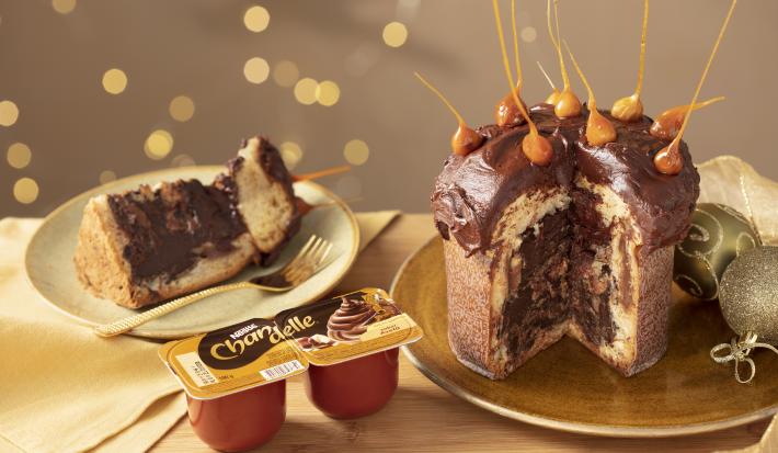 Fotografia em tons de amarelo em uma bancada de madeira clara, um pano amarelo e decoração natalina de fundo. Ao centro, um prato dourado com o panettone de avelã recheado sem um pedaço. Ao lado, um prato com a fatia do panettone.