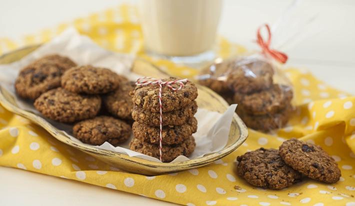 Fotografia em tons de branco e amarelo de uma bancada branca com um paninho amarelo com bolinhas brancas, sobre ele cookies. Ao fundo um copo de vidro.