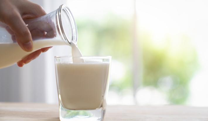 Fotografia de uma mão colocando uma bebida de iogurte com leite e cereal em um copo de vidro, que está sobre uma mesa de madeira em tom claro.