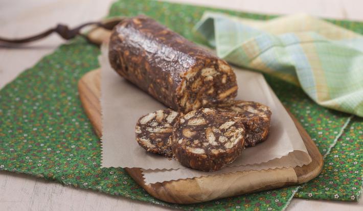 Fotografia em tons de verde em uma bancada de madeira com um pano verde florido, uma tábua de madeira com um papel bege e salaminho de chocolate.