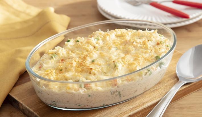 Foto da Receita de Arroz de Forno Cremoso com Sobras de Frango e Seleta de Legumes. Observa-se um refratário de vidro oval com o arroz gratinado e, ao lado, uma colher de servir de inox.