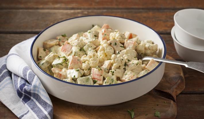 Fotografia em tons de marrom, branco e azul de uma bancada de madeira marrom com uma tábua de madeira, sobre ela um paninho branco xadrez com um recipiente redondo com a salada de legumes  e uma colher. Ao lado dois recipientes brancos.