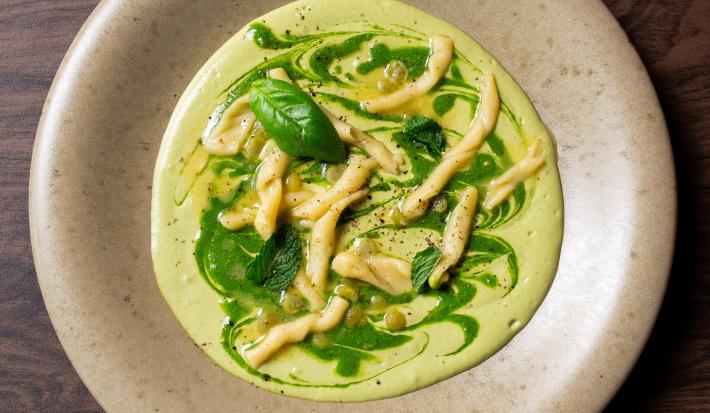 Fotografia em tons de cinza com um prato cinza e fundo ao centro. Dentro do prato existe uma receita de macarrão trofie com molho de abobrinha e ervilhas coberto com pesto de ervas.