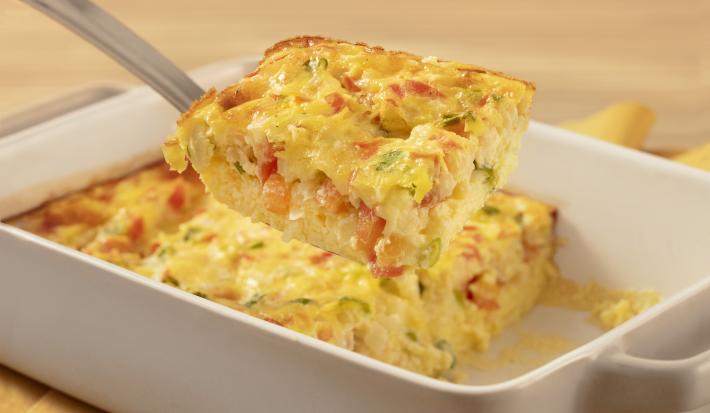 Fotografia em tons de branco e amarelo de uma bancada de madeira clara com um paninho amarelo sobre ele um recipiente quadrado branco com o omelete de forno e uma espátula retirando um pedaço de omelete.