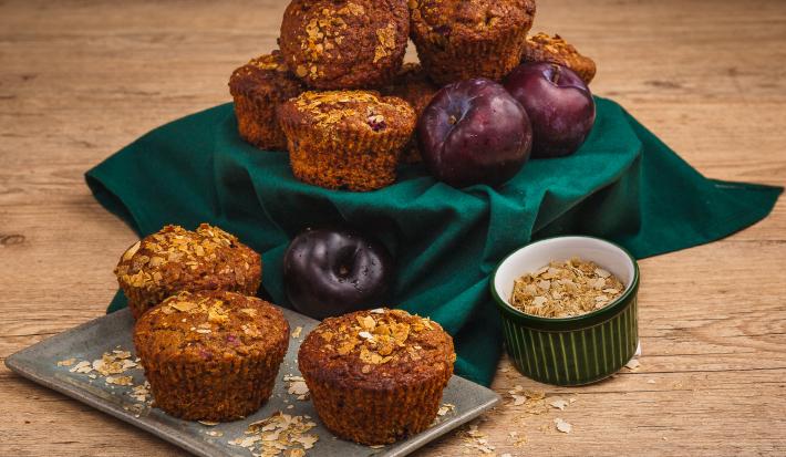 Fotografia em tons de verde em uma bancada de madeira clara, um pano verde, um recipiente retangular com os muffins de ameixa e cereais em cima. Ao lado, um potinho com cereais. Ao fundo, um suporte com o pano verde e mais muffins e ameixas.