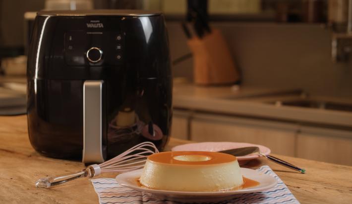 Imagem de um pudim de leite condensado Moça sobre um prato branco, ao lado de um fouet e uma air fryer preta, em uma bancada de cozinha.