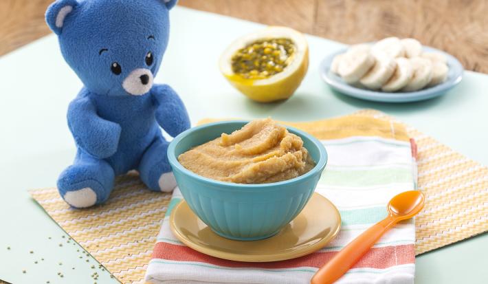 Fotografia em tons de azul em uma bancada de madeira com paninho listrado em azul e vermelho e um potinho azul com a delicinha de maracujá sobre um pratinho laranja, ao lado um ursinho de pelúcia azul. Ao fundo, um pedaço de maracujá e fatias de banana.