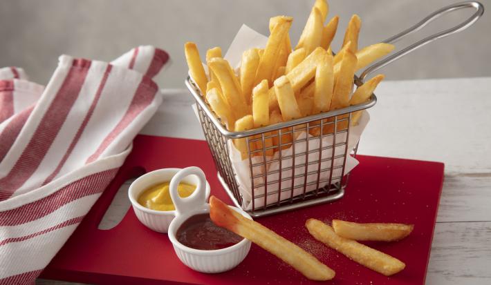 Fotografia em tons de vermelho de uma bancada branca, ao centro uma tábua vermelha com uma cestinha com batatas fritas dentro. Na frente um potinho com ketchup e um com mostarda.