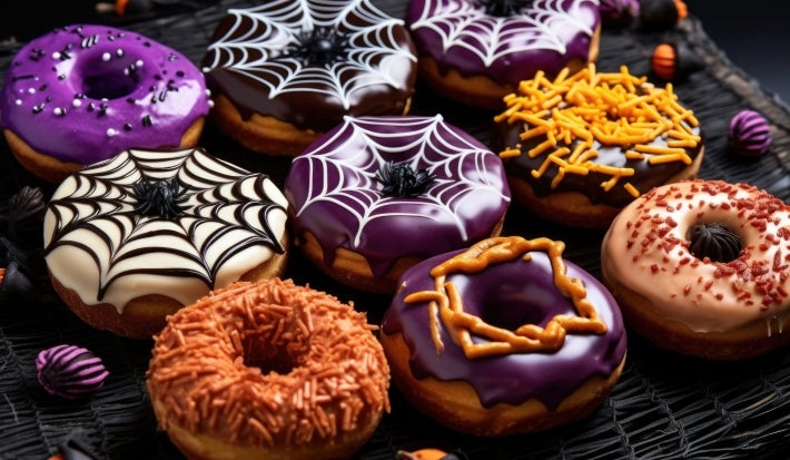 Fotografia em tons escuros e coloridos com vários donuts ao centro. Os donuts são enfeitados com temas de halloween como: miconhas e teias de aranha feitas nas cores roxo, laranja, preto e branco.