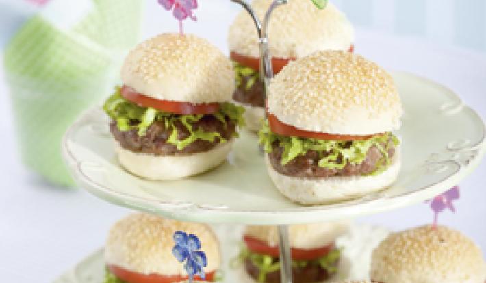 Fotografia em tons de verde em uma mesa branca com um pedestal enfeitado com borboletas de papel de pratos brancos com os hamburguinhos colocados nele. Ao fundo, um copo com guardanapos de papel verde e rosa.