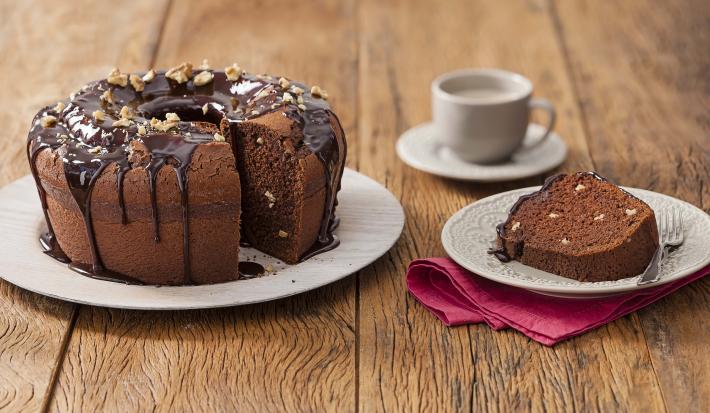 Fotografia em tons de marrom em uma mesa de madeira com um prato branco grande e o bolo de chocolate e nozes e ao lado um prato branco pequeno com uma fatia do bolo. Ao fundo, um pires e uma xícara branca.