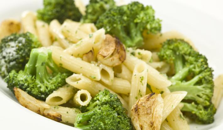 Fotografia de uma receita de penne com brócolis, um pouco de azeitona e alho assado dentro de um refratário fundo branco sobre uma mesa de cor branca.