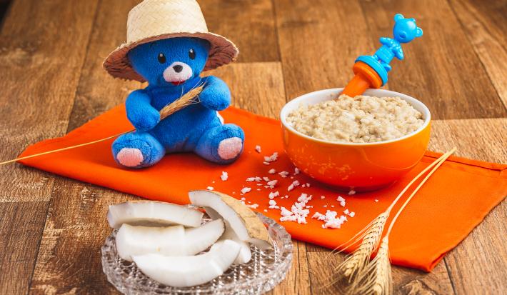 Fotografia em tons de laranja e azul de uma bancada de madeira com um paninho laranja e um pote laranja com a papinha. Na frente, fatias de coco seco decorando e uma pelúcia azul com chapéu de palha.