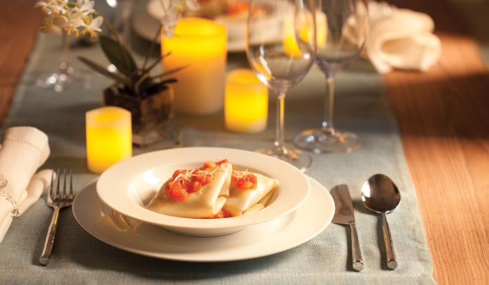 Fotografia em tons de amarelo em uma mesa de madeira e uma toalha de mesa cinza. Um sousplat branco e um prato fundo branco com a sofioli de shimeji ao molho de tomates. Velas amarelas, taças de vidro, arranjo pequeno de flor e talheres compondo a mesa.