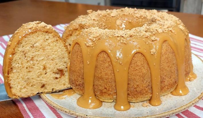 Foto em tons de caramelo da receita de bolo de paçoca com leite ninho servida em uma porção inteira com um pedaço cortado sobre uma base de cerâmica em cima de um paninho listrado rosa
