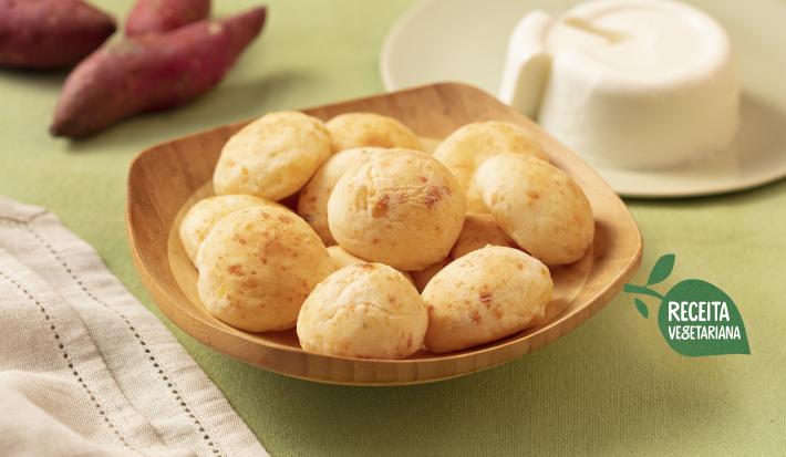 Fotografia em tons de verde e branco de uma bancada de verde clara com um recipiente de madeira clara com pães de queijo. Ao fundo um prato branco com o queijo minas e duas batatas-doces. Na frente um paninho branco.