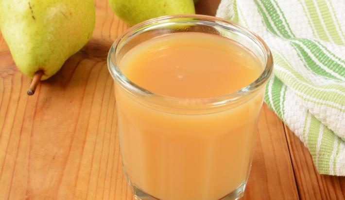 Fotografia em tons de marrom, laranja, branco e verde de uma bancada de madeira marrom vista de cima, contém um copo transparente com suco de pera e maçã. Ao fundo duas peras e ao lado um pano branco e verde