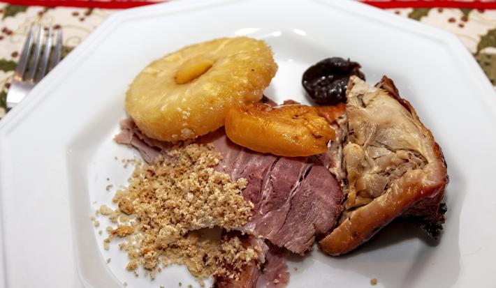 Fotografia vista de cima de um corte de carne, duas fatias de lombo, uma fatia de abacaxi assado e duas frutas assadas em um prato branco. Ao lado do prato, um garfo que está sobre uma toalha de mesa desenhada, na cor branco e verde.