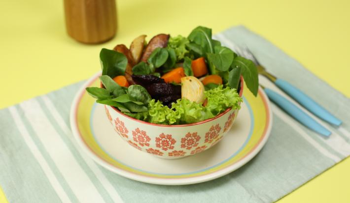 Fotografia em tons de amarelo em uma bancada de madeira amarela, um jogo americano verde claro listrado, um prato branco com listas coloridas, um potinho branco com flores e dentro uma salada de legumes.