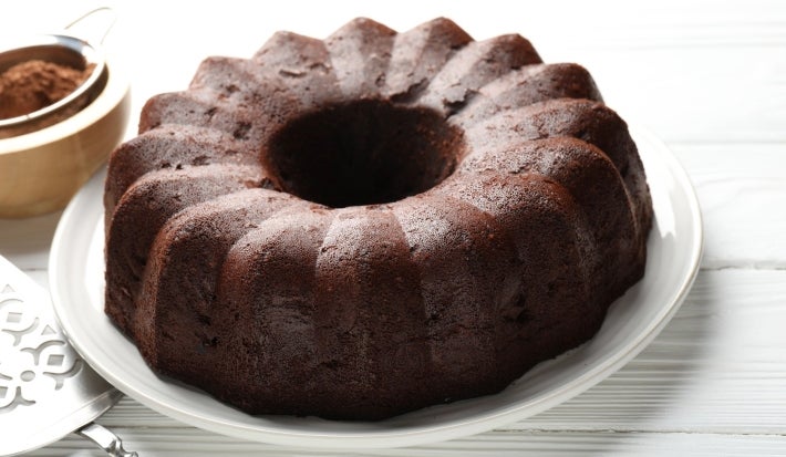 Em cima de uma mesa, bolo de chocolate com aparência escura e densa com úmida e rica em cacau. A textura da superfície é rústica e com coloração marrom intensa. Esse bolo ainda está inteiro, sem cortes ou fatias separadas.
