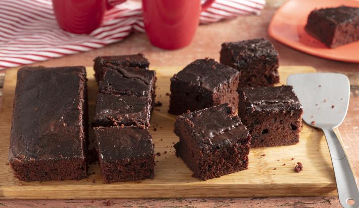 Fotografia em tons de marrom e vermelho de uma bancada de madeira, ao centro uma tábua com fatias de bolo de chocolate. Ao fundo 2 xícaras e um prato vermelho com uma fatia de bolo.