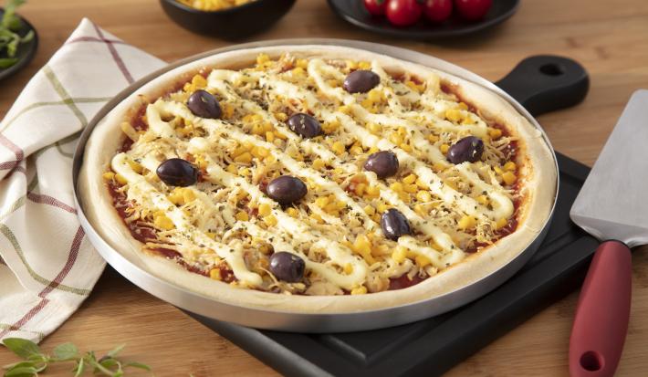 Imagem de uma pizza de frango com catupiry, milho e azeitonas sobre uma forma de alumínio.