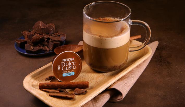 Fotografia em tons de marrom em uma bancada de madeira marrom escura, um recipiente oval de madeira clara, um pano bege, uma xícara de vidro com a bebida de café ao leite desnatado com chocolate e canela dentro dela. Ao fundo, pedaços de chocolate.