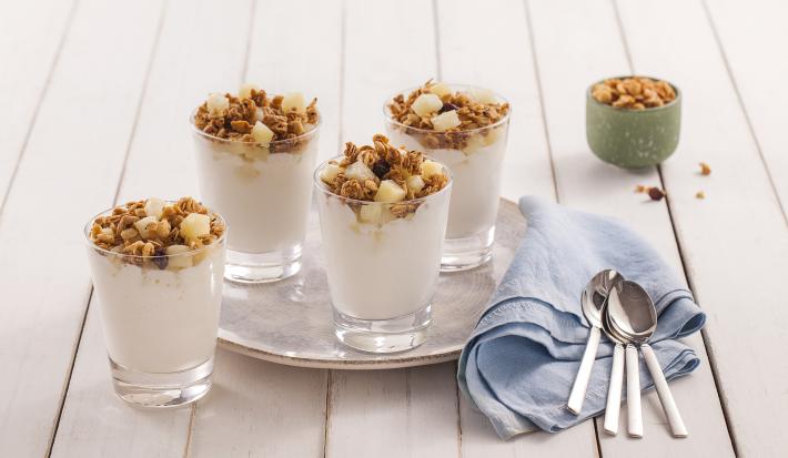 Fotografia em tons de azul em uma bancada de madeira clara, um paninho azul, um prato redondo raso grande com copinhos de vidro com a sobremesa gelada de abacaxi com granola. Ao fundo, um potinho verde com granola.