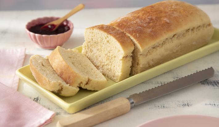 Receita de Pão de Leite Moça. Observa-se um pão cortado com três fatias sob uma forma retangular verde clara. Ao lado, uma faca de pão e do outro, um potinho com geleia.