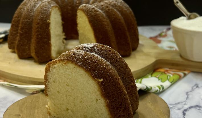 Foto da receita de Bolo Fácil de Iogurte, com uma fatia mais próxima, sobre uma tábua de madeira e o restante do bolo ao fundo, servido também numa tábua, ao lado de um pote com açúcar.