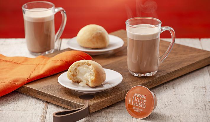 fotografia em tons de laranja e marrom de uma bancada cinza vista de frente, contém uma tábua de madeira com dois pratinhos redondos e brancos com pão de batata com requeijão em cada um, uma xicara transparente com mochaccino canela e um pano laranja.