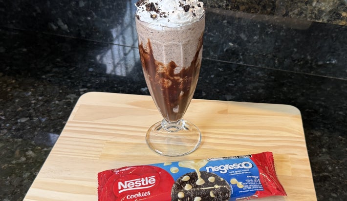 Foto da receita de Milkshake Cookies and Cream, servido em um copo alto decorado com calda de chocolate, com chantilly em cima, sobre uma tábua de madeira com um pacote de Cookies Negresco