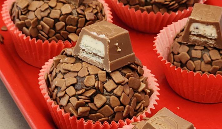 Fotografia de brigadeiros cuidadosamente enrolados em granulado e decorados com pedaços de KitKat, apresentados em forminhas vermelhas sobre um prato da mesma cor.