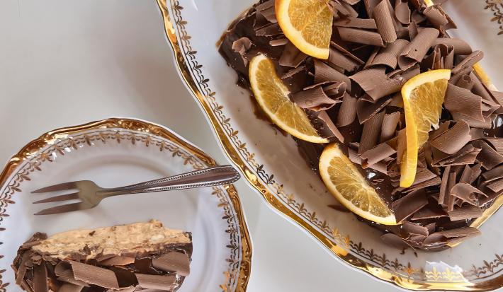 Fotografia em tons de brano com uma travessa de vidro ao centro. Em cima da travessa existe uma receita de Choco Torta coberta com raspas de chocolate ao leite e pedaços de laranja