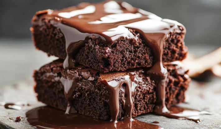 Um pedaço de bolo de chocolate empilhado sobre outro, ambos com uma cobertura generosa de calda de chocolate derretido escorrendo pelas laterais. A textura é úmida e densa, com aparência extremamente saborosa.