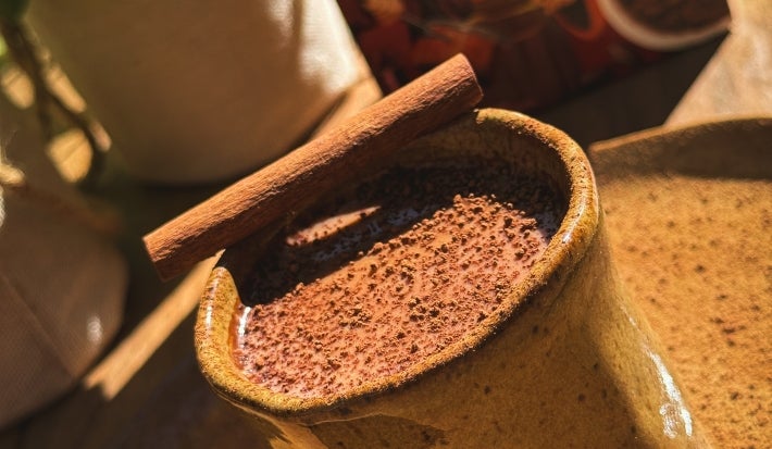 Fotografia em tom claro sob o sol de um prato grande e raso de tom marrom sobre uma mesa de madeira. Apoiado no prato tem um copo de vidro no memso tom com chocolate quente e um pau de cnaela na borda do copo.