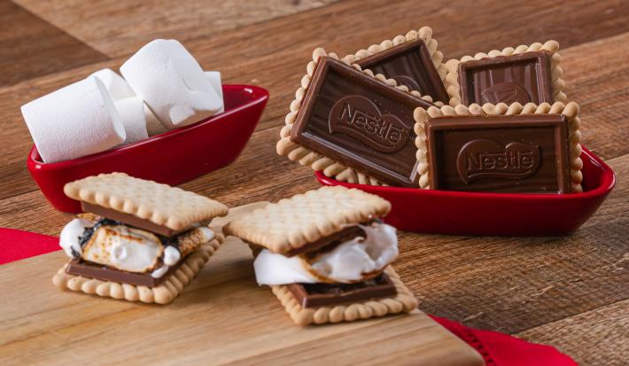 Fotografia em tons de vermelho com vários recipientes vermelhos ao centro. Dentro de um dos recipientes, existe dois biscoitos Choco Biscuit recheados com marsmallow. Ao lado existe marshmallows e biscoitos Choco Biscuit NESTLÉ