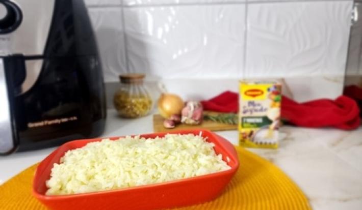 Foto da receita de arroz na airfryer servida em uma travessa vermelha com uma airfryer ao fundo junto com uma embalagem de maggi meu segredo, cebolas e um pano vermelho