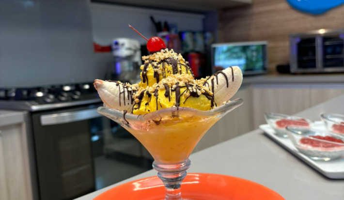Fotografia de uma taça de vidro com banana split com calda de chocolate, farofa de castanha e sorvete de abacate.