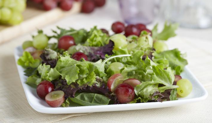 Fotografia de um refratário branco quadrado raso com uma salada de alface, agrião salsão e uva itália com molho de iogurte. O recipiente está apoiado em uma toalha de mesa em tom de bege. Ao fundo tem dois cachos de uva, sendo uvas verdes e uvas roxas.