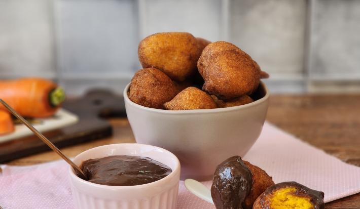 Foto da receita de bolinho de chuva servida em diversas porções em um bowl de cerâmica bege sobre um paninho rosa com um recipiente com a calda a frente. Ao fundo há uma tábua com cenouras em cima de uma mesa de madeira