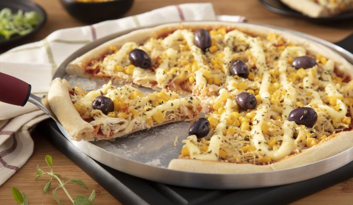 Fotografia em tons amarelo em uma bancada de madeira, uma tábua de alumínio redonda com a pizza de frango com catupiry em cima dela. Ao lado, potinhos com milho, azeitonas e um prato com uma fatia da pizza.
