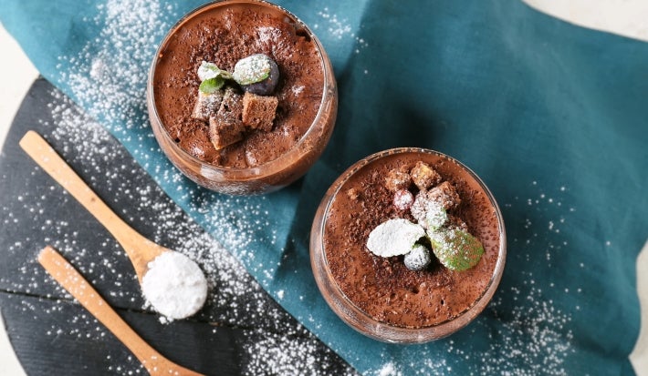 Fotografia vista de cima de dois copos de vidro decorativos sobre um peno de tom azul dobrado, com açúcar de confeiteiro polvilhado por cima do pano e da sobremesa. Dentro do copo tem a sobremesa, finalizada com ganache, uva e cacau em pó.