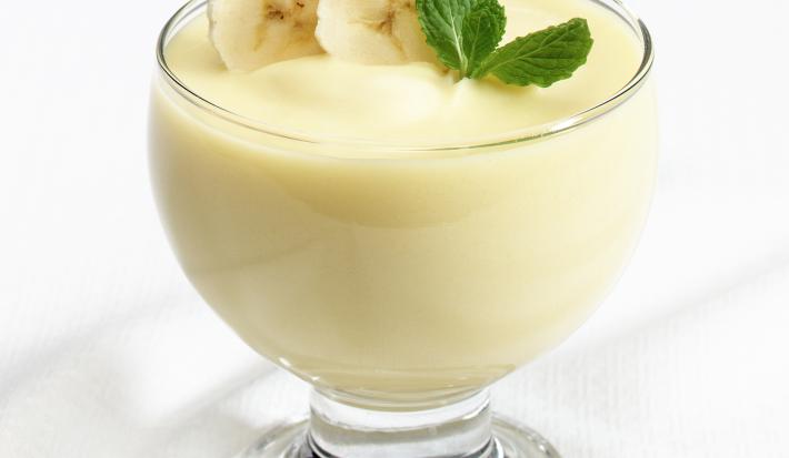 Fotografia de um flan de leite MOÇA e banana dentro de um recipiente de vidro com rodelas de banana e folhas de hortelã. A sobremesa está sobre uma mesa branca.