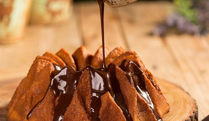 Fotografia em tons de marrom em uma bancada de madeira e ao centro uma tábua de tronco de árvore com o bolo de cenoura em cima e uma mão segurando um potinho despejando a calda de chocolate por cima do bolo.